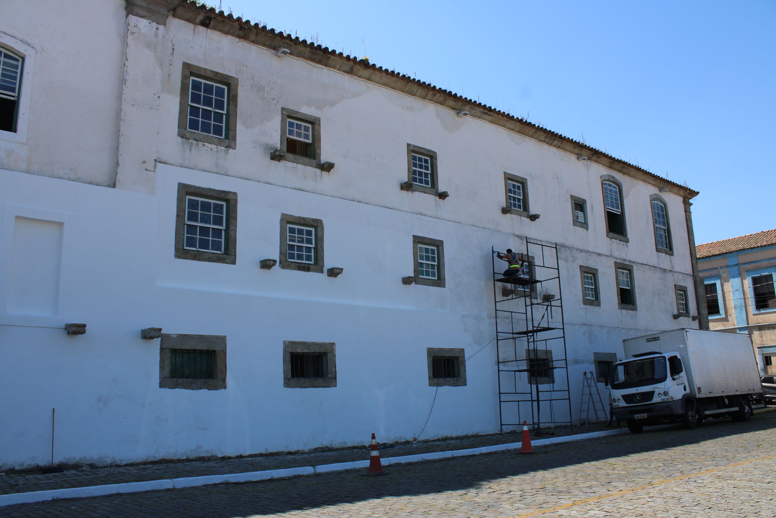 Imagem de uma parede de edifício em reforma, mostrando um trabalhador em andaime e veículo de carga ao lado em cenário urbano. Museu de Arqueologia e Etnologia (MAE) da UFPR ganha nova pintura