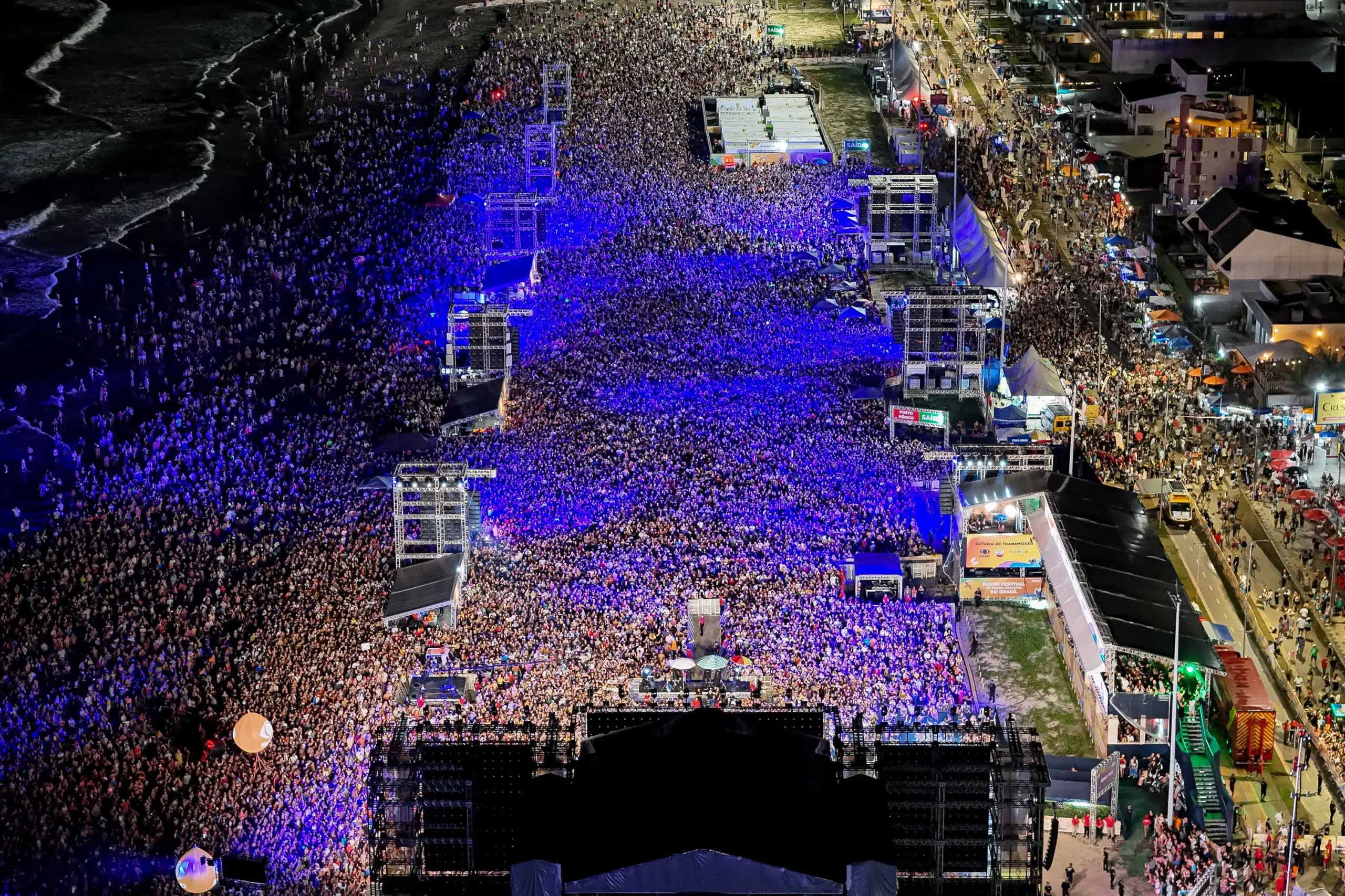 Grande evento noturno à beira-mar com multidão animada sob luzes vibrantes, capturando a energia de um festival de música. Shows do Verão Maior reúnem 1 milhão de pessoas; número ultrapassa temporada anterior