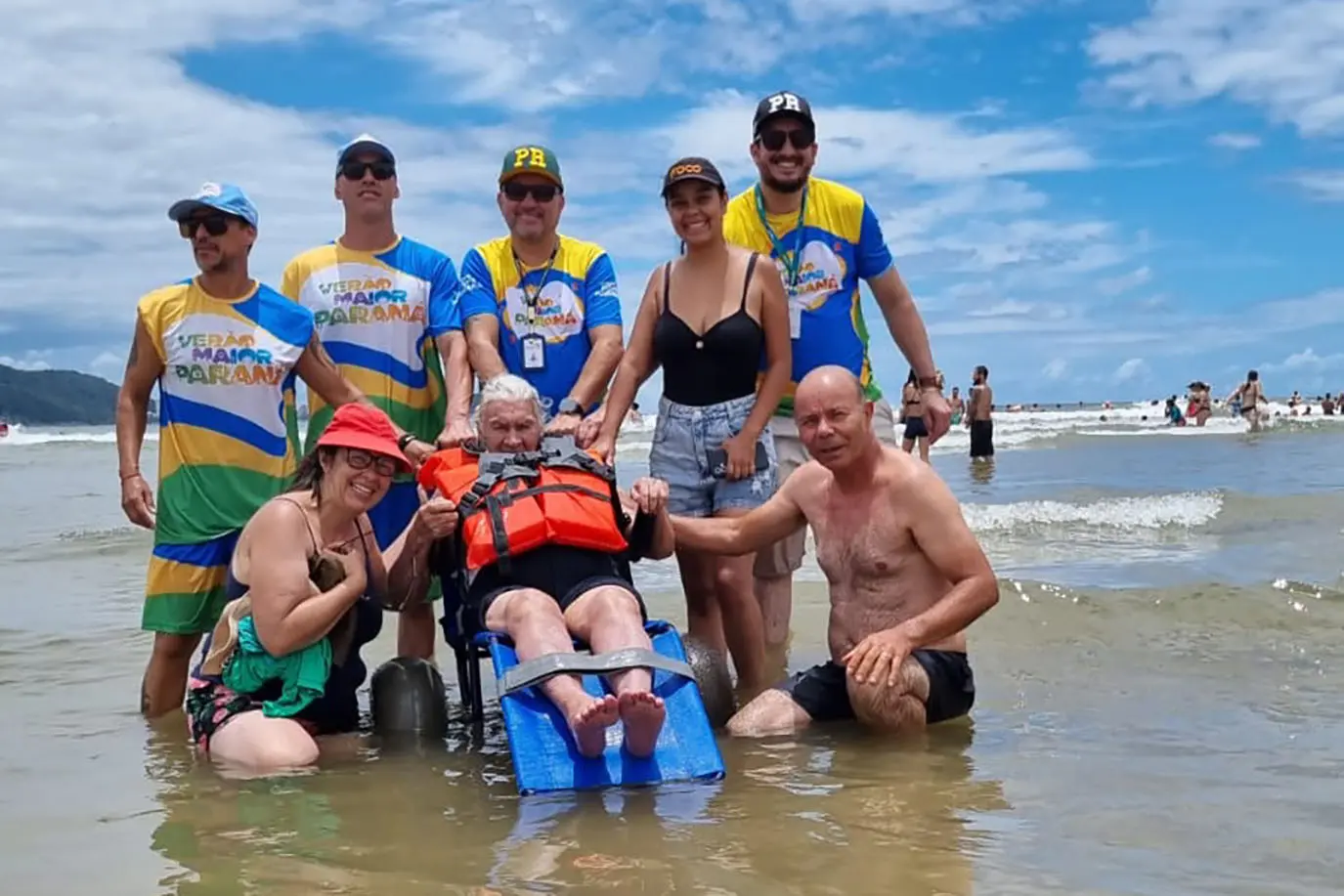 Grupo de pessoas sorrindo na praia com um idoso em cadeira de rodas adaptada para o mar, promovendo a inclusão e acessibilidade para todos. Com 1.068 atendimentos, cadeiras anfíbias batem recorde de utilização no Verão Maior