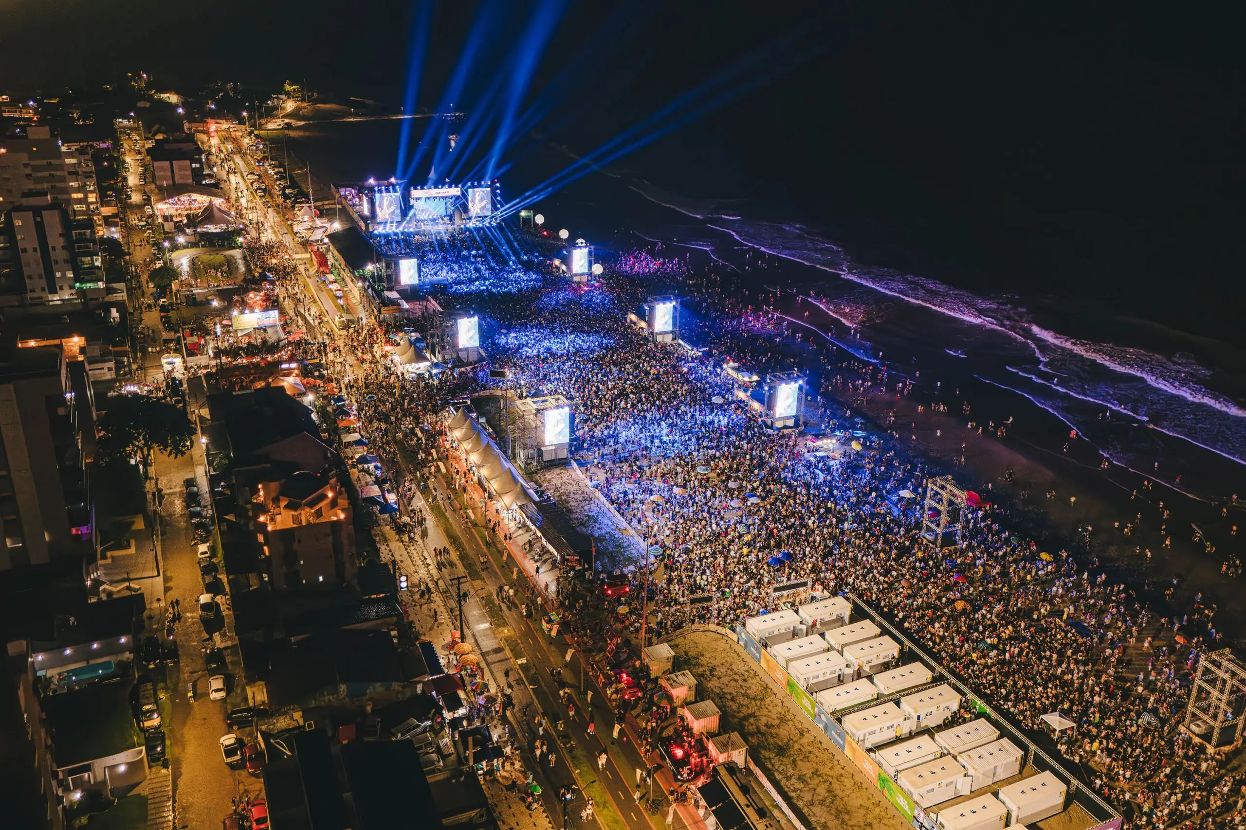 Vista aérea de uma festa à beira-mar durante a noite, com milhares de pessoas, shows ao vivo e luzes brilhantes. Ótimo exemplo de festividade popular.Verão Maior Paraná: shows têm público superior a 200 mil pessoas em todos os finais de semana