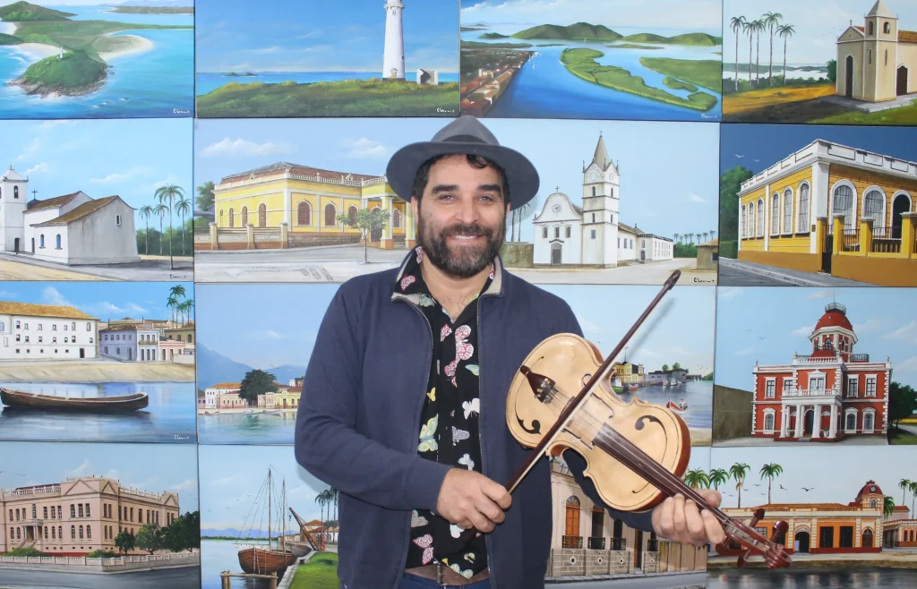 Homem sorridente segurando um instrumento musical em frente a um fundo com várias pinturas de paisagens e edifícios, representando a cultura brasileira. Baile do Fandango Caiçara acontece neste sábado, na Associação Mandicuera
