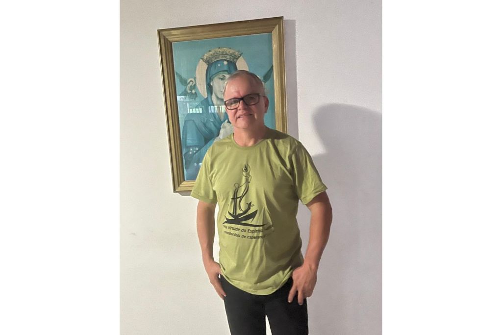 Homem com camiseta verde e óculos, posando em frente a uma pintura na parede. A camiseta exibe um desenho de âncora com uma frase. Renovação Carismática Católica promove o Cristoval 2025 no dia 4 de março
