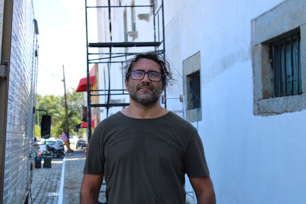 Homem com óculos e cabelo desgrenhado em um ambiente urbano, próximo a um muro e andaimes, retratando uma cena descontraída e natural. Museu de Arqueologia e Etnologia (MAE) da UFPR ganha nova pintura
