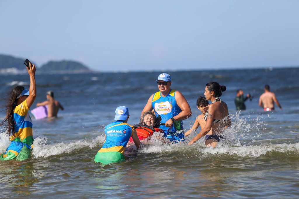 Grupo de pessoas se divertindo na praia, incluindo crianças brincando na água com salva-vidas. Um momento alegre e seguro no litoral. Com 1.068 atendimentos, cadeiras anfíbias batem recorde de utilização no Verão Maior
