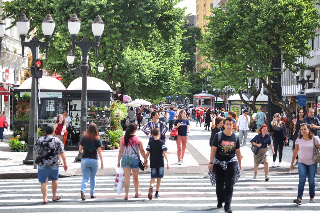 Imagem de uma movimentada rua urbana em um dia ensolarado, com muitas pessoas atravessando a faixa de pedestres e lojas ao fundo, destacando a vitalidade da cidade. Comércio do Paraná cresce 5,3% em 2024 e supera média nacional
