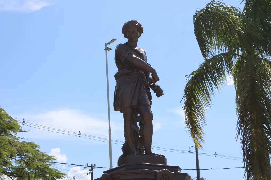 Estátua de bronze de um jovem segurando flores, cercada por palmeiras, sob um céu azul claro. Localização ao ar livre, ideal para fotos. Chafariz do Outono: Conheça a importância histórica do monumento de Paranaguá