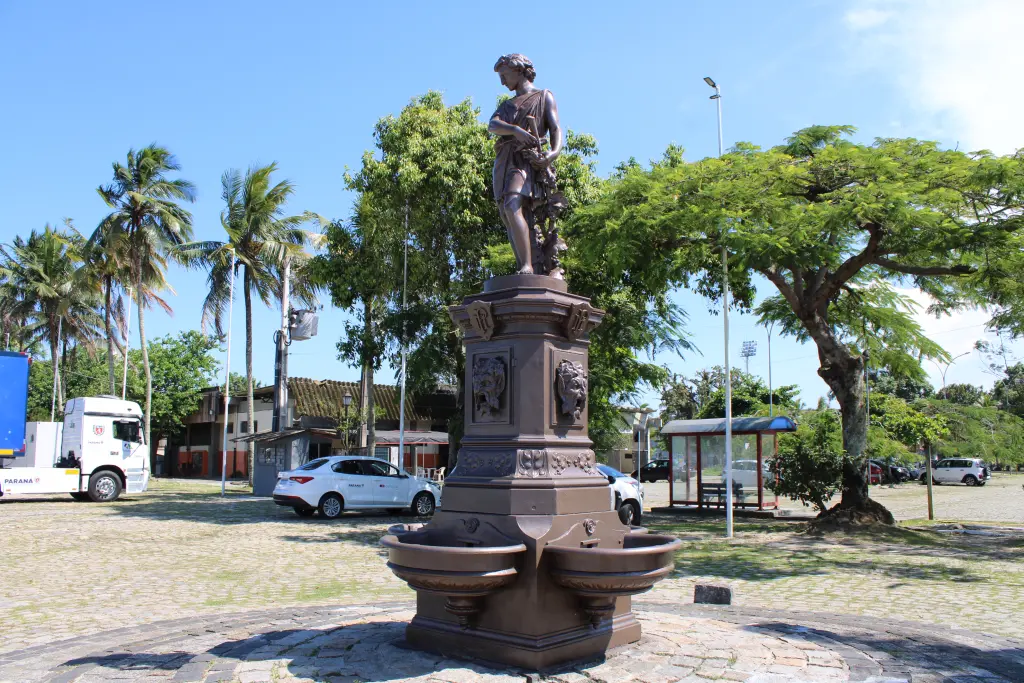 Estátua de bronze com um jovem segurando um objeto, rodeada por palmeiras e edifícios urbanos, destacando a beleza da arte pública em ambientes naturais. Chafariz do Outono: Conheça a importância histórica do monumento de Paranaguá