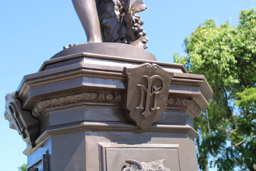 Detalhe da base de uma estátua com um emblema em relevo, rodeada por vegetação verde e sob um céu azul claro, representando arte e cultura. Chafariz do Outono: Conheça a importância histórica do monumento de Paranaguá