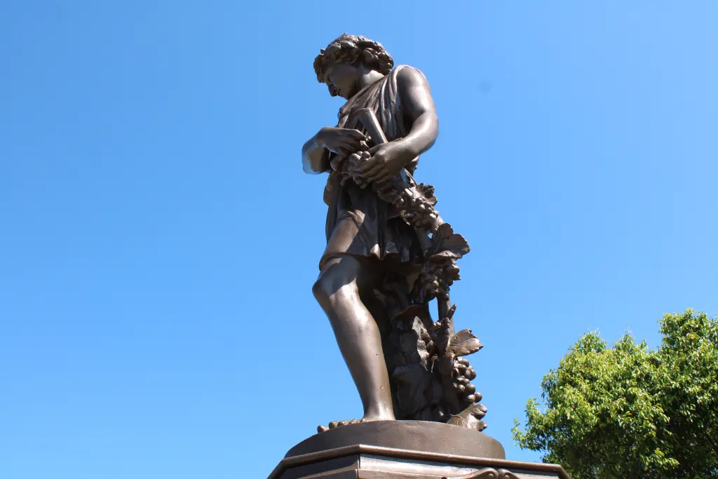 Estátua de um jovem segurando uvas, simbolizando a fertilidade e a natureza, em um dia ensolarado com céu azul. Chafariz do Outono: Conheça a importância histórica do monumento de Paranaguá