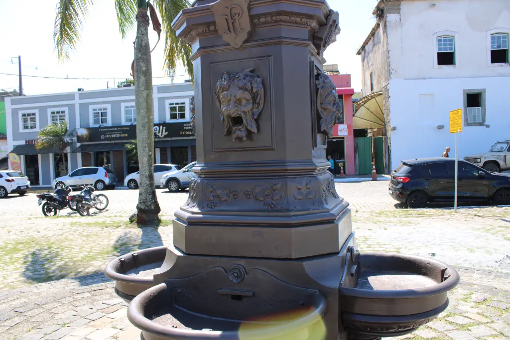 Fonte decorativa com detalhes em forma de rostos antropomórficos, localizada em área urbana com veículos e edifícios ao fundo. Chafariz do Outono: Conheça a importância histórica do monumento de Paranaguá