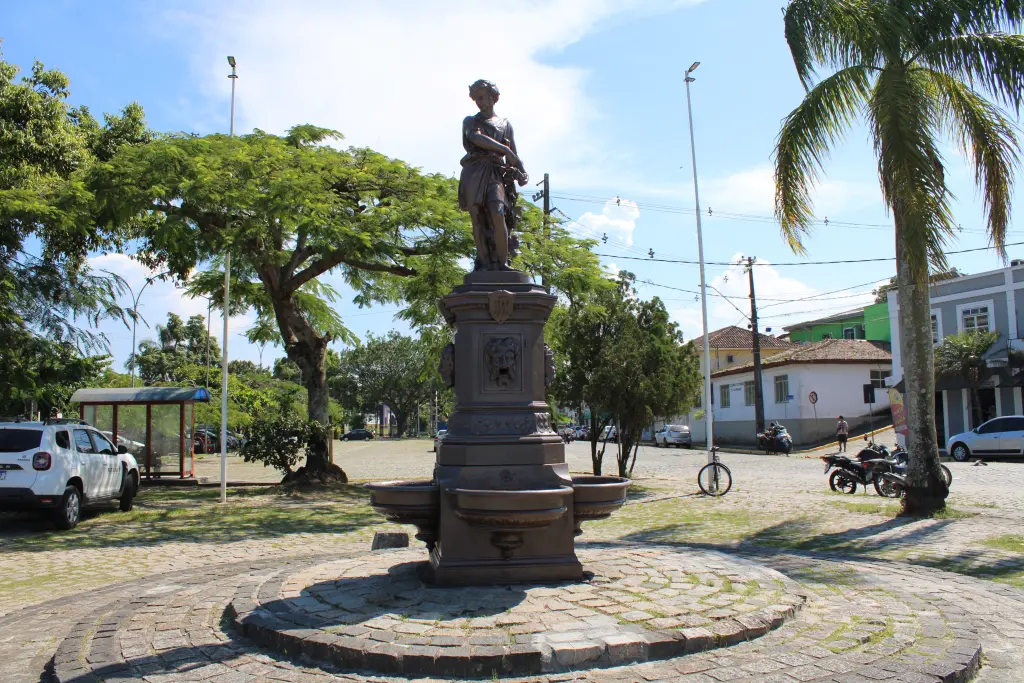 Estátua em praça urbana, cercada por árvores e lojas. Imagem destaca a beleza arquitetônica e a natureza ao redor. Chafariz do Outono: Conheça a importância histórica do monumento de Paranaguá 