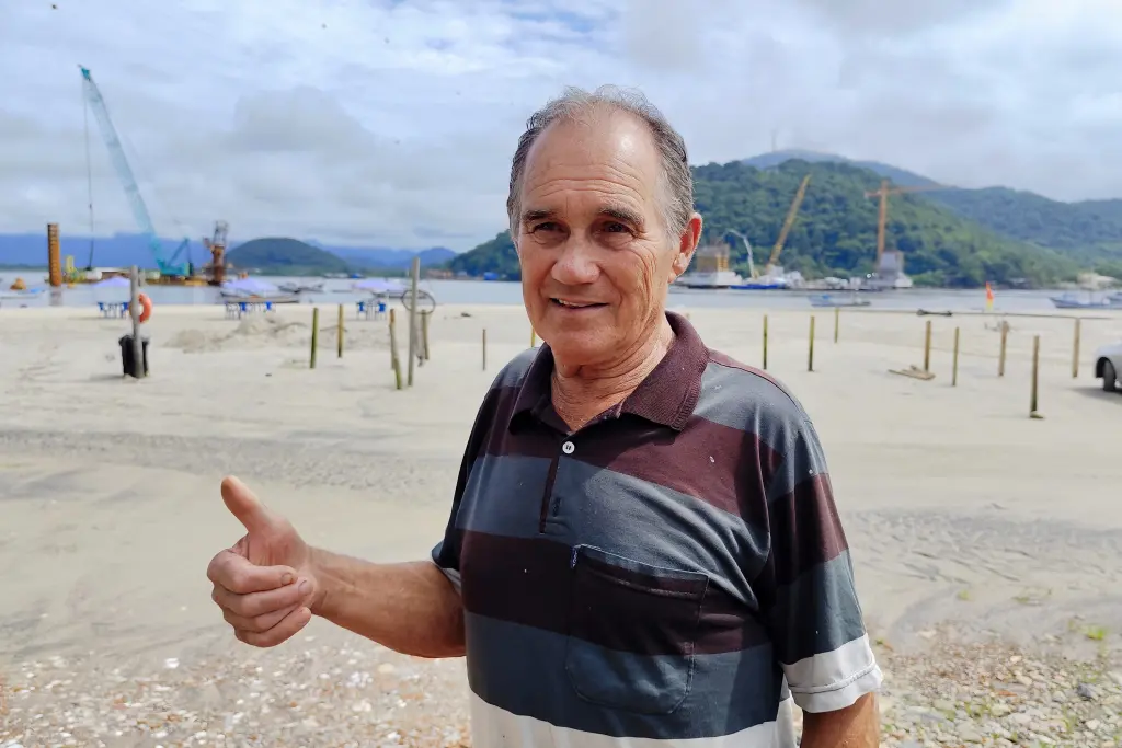 Homem sorridente com gesto positivo em praia, ao fundo cenário de construção e montanhas. Ideal para promoção de turismo e atividades ao ar livre. Moradores e comerciantes comemoram progresso das obras da Ponte de Guaratuba
