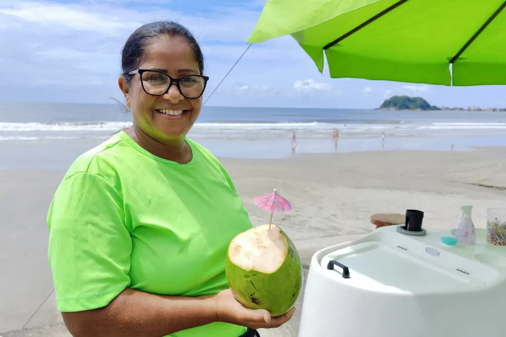 Mulher sorridente de camiseta verde segurando uma água de coco com guarda-chuva na praia, em um dia ensolarado. Moradores e comerciantes comemoram progresso das obras da Ponte de Guaratuba
