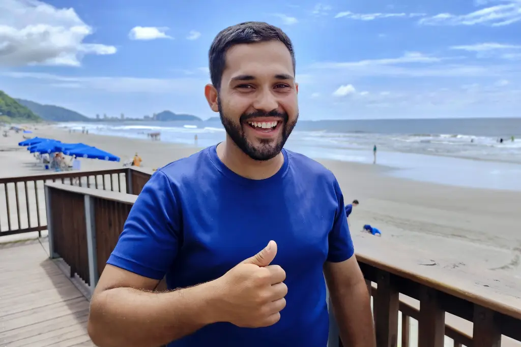 Homem sorridente mostrando o polegar para cima na praia, com céu azul e ondas ao fundo, ideal para conteúdo sobre diversão e relaxamento na costa. Moradores e comerciantes comemoram progresso das obras da Ponte de Guaratuba
