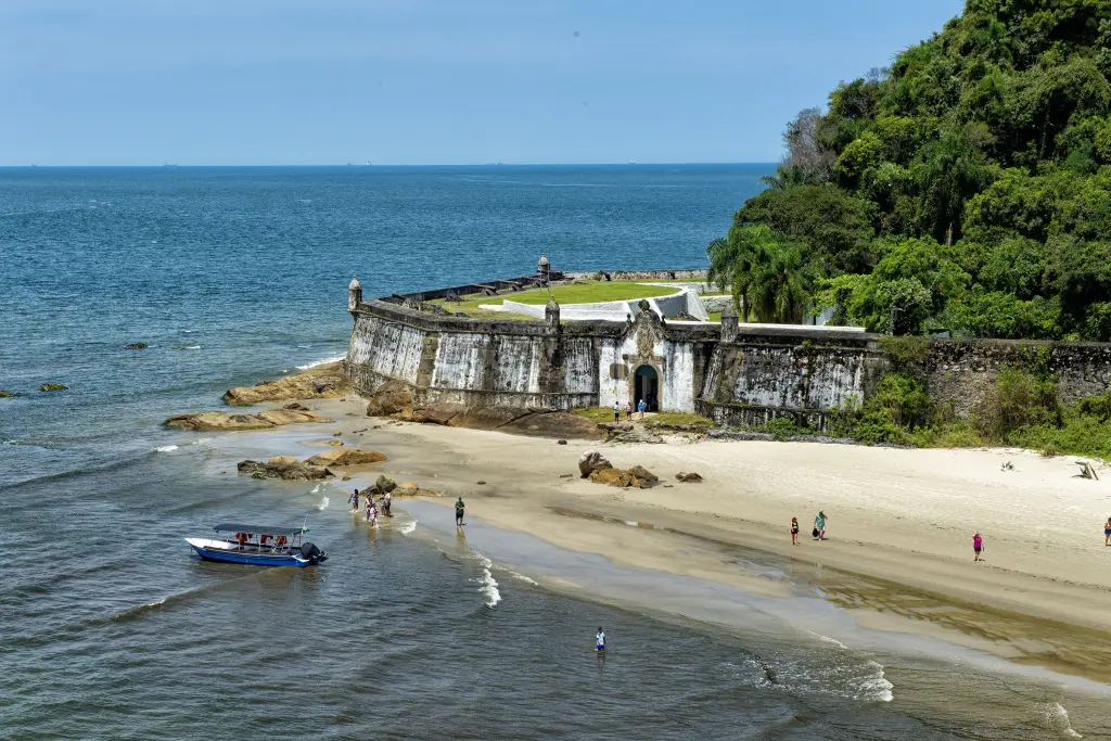 Vista do forte à beira-mar, próximo à praia, com pessoas relaxando e um barco na água azul, representando um cenário lindo e histórico. Carnaval na Ilha do Mel: 10 orientações para curtir o feriado na Unidade de Conservação
