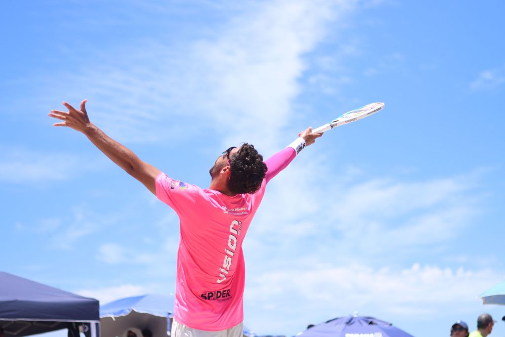 Atleta praticando beach tennis na praia, com raquete e camiseta rosa sob um céu azul claro. Com 128 atletas, circuito mundial de beach tennis chega a Caiobá nesta quarta-feira
