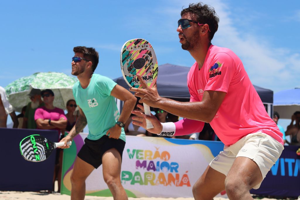 Dois jogadores de beach tennis em ação na praia, destacando-se em um evento de verão no Paraná. Os atletas usam roupas coloridas e interagem com o público animado.