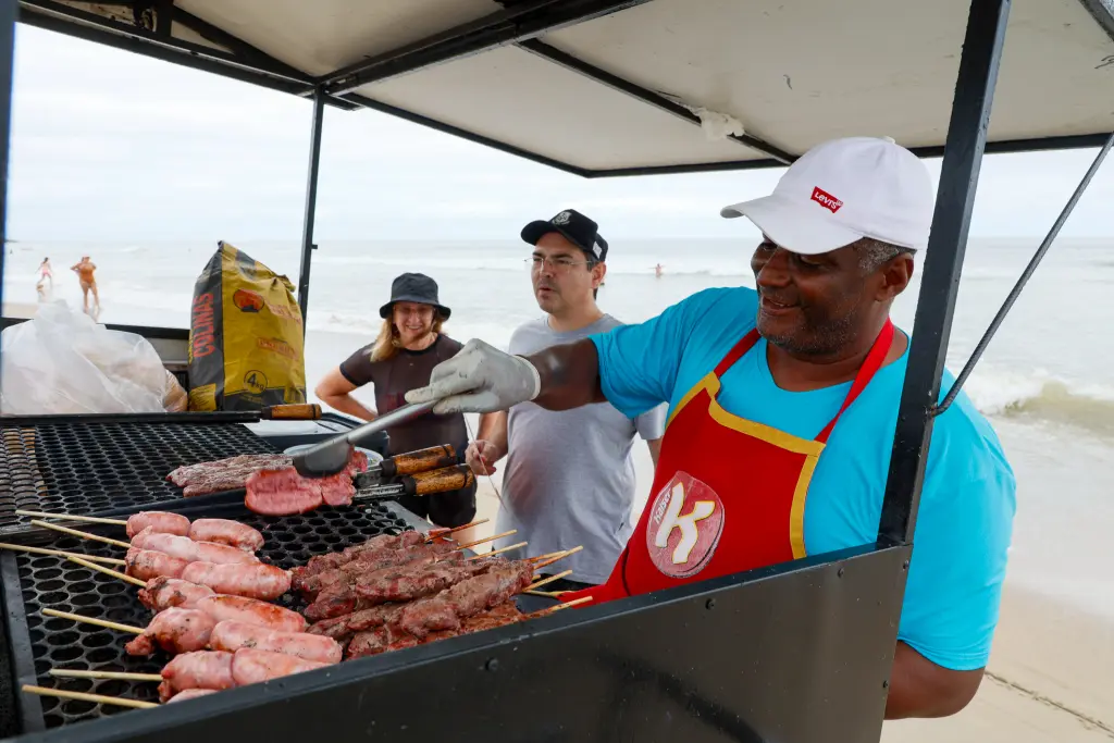 Um vendedor de churrasco prepara carnes em uma grelha na praia, enquanto clientes observam. O ambiente é ensolarado e relaxante. Verão Maior Paraná termina com mais de 20% de crescimento nas vendas do comércio do Litoral
