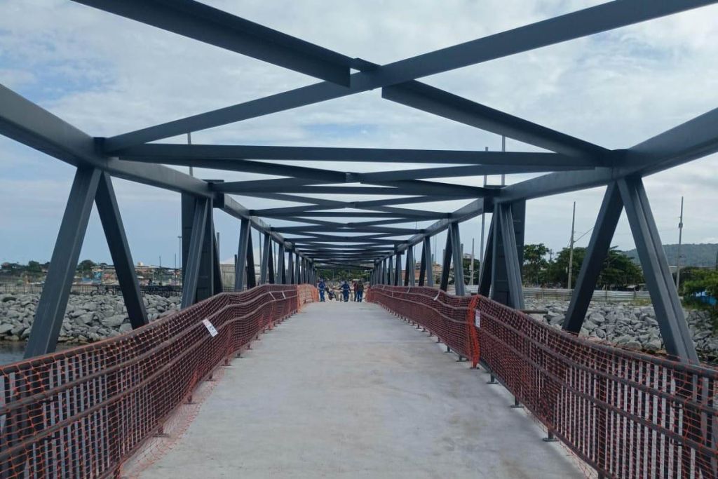 Imagem de uma ponte moderna com estrutura metálica e passarela em obra, vista sob um céu nublado. Destaca-se a união de design e funcionalidade na arquitetura urbana. Peça importante na revitalização da orla, passarela do Rio Matinhos é entregue pelo IAT
