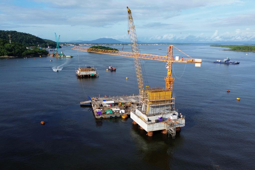 Imagem de uma plataforma em construção no mar, destacando um guindaste amarelo e atividade portuária ao fundo, refletindo novos empreendimentos e infraestrutura. Com avanço no trecho estaiado, obras da Ponte de Guaratuba chegam a 38,8% de execução
