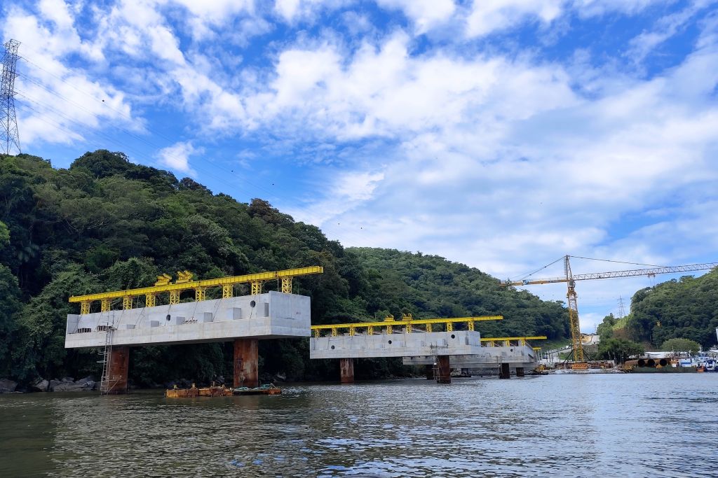 Imagem de uma construção em andamento na água, com estruturas inacabadas e guindaste ao fundo, cercada por vegetação. O céu está parcialmente nublado. Com avanço no trecho estaiado, obras da Ponte de Guaratuba chegam a 38,8% de execução
