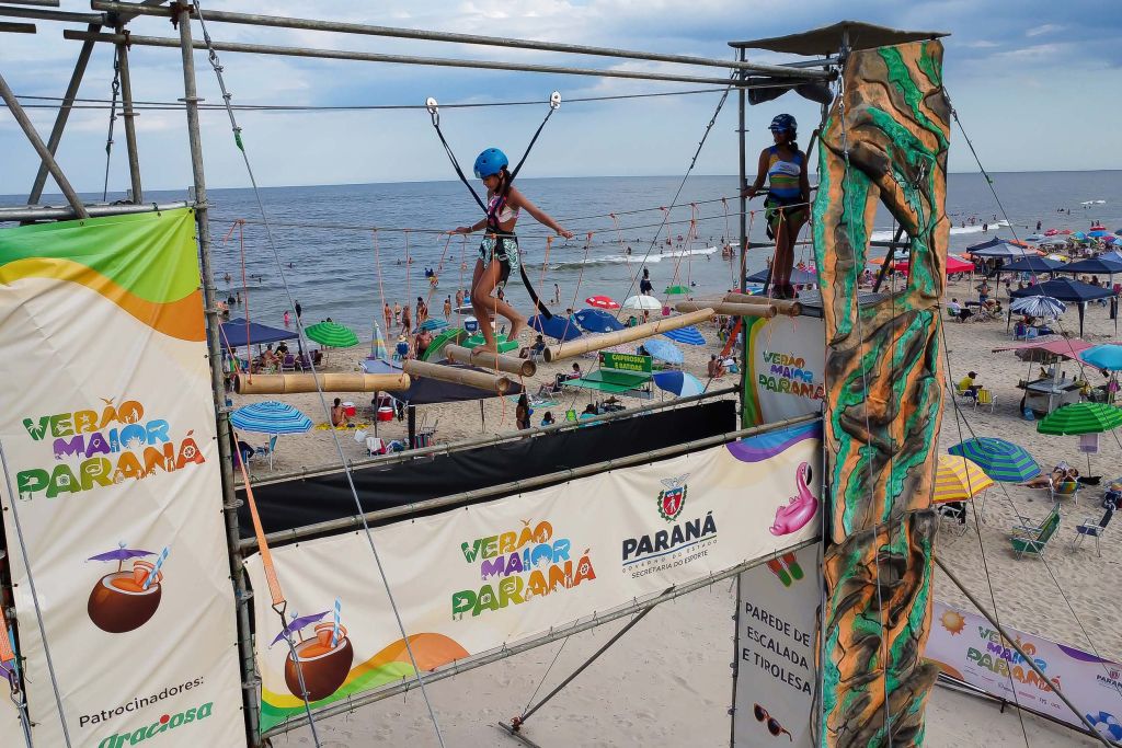 Crianças se divertindo em atividade de lazer na praia durante o verão no Paraná, com estrutura de cordas e balanços em segundo plano. Programação e atividades do Verão Maior têm 85% de aprovação entre paranaenses. Programação e atividades do Verão Maior têm 85% de aprovação entre paranaenses
