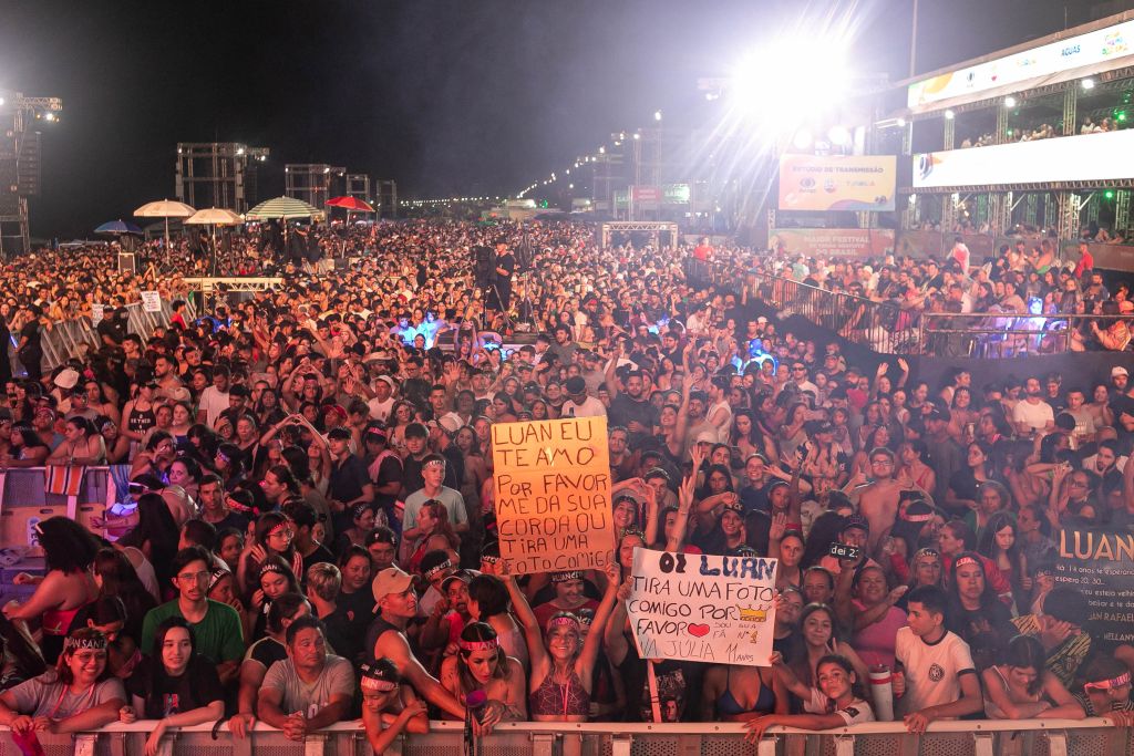 Multidão em show ao ar livre, com fãs segurando cartazes pedindo fotos e declarando amor ao cantor Luan. A energia é intensa e vibrante, marcada por luzes e animação. Shows do Verão Maior reúnem 1 milhão de pessoas; número ultrapassa temporada anterior