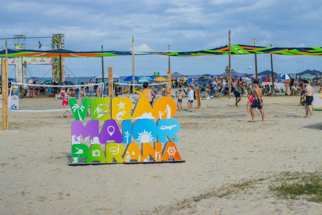 Praia com pessoas se divertindo durante o verão, com cartaz colorido "Verão Maior Paraná" e barracas ao fundo. Um dia ensolarado perfeito para lazer. Postos fixos do Verão Maior Paraná encerram atividades com 2,4 milhões de atendimentos
