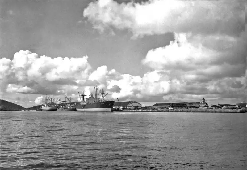 Portos do Paraná: 90 anos de história impulsionando o desenvolvimento de Paranaguá 4 Imagem em preto e branco de um porto com navios atracados e um céu nublado, destacando a atividade portuária e a paisagem ao redor. Portos do Paraná: 90 anos de história impulsionando o desenvolvimento de Paranaguá