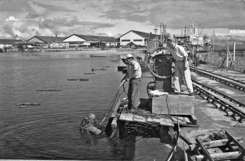 Portos do Paraná: 90 anos de história impulsionando o desenvolvimento de Paranaguá 6 Imagem em preto e branco mostrando mergulhadores trabalhando em um cais, com homens ao redor e barcos ao fundo. A cena retrata atividades de mergulho em ambiente portuário. Portos do Paraná: 90 anos de história impulsionando o desenvolvimento de Paranaguá