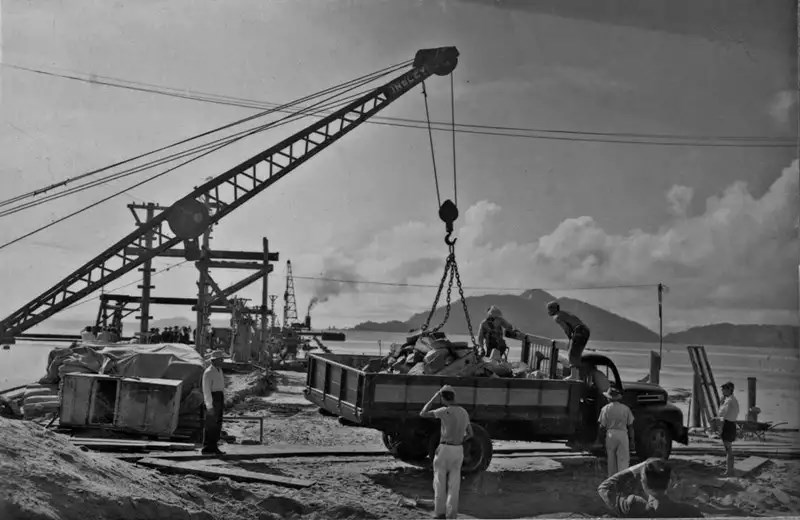 Portos do Paraná: 90 anos de história impulsionando o desenvolvimento de Paranaguá 3 Imagem histórica de um canteiro de obras com um guindaste e trabalhadores carregando material em um caminhão, em um cenário de praia. Portos do Paraná: 90 anos de história impulsionando o desenvolvimento de Paranaguá