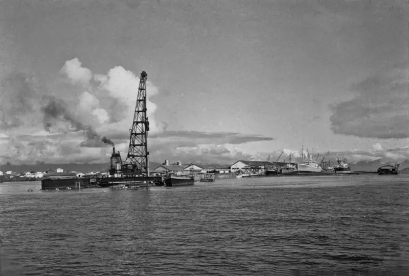 Portos do Paraná: 90 anos de história impulsionando o desenvolvimento de Paranaguá 5 Vista histórica de uma plataforma de perfuração e embarcações em um porto, com céu nublado e reflexo na água. Imagem em preto e branco que retrata a indústria navais e de exploração. Portos do Paraná: 90 anos de história impulsionando o desenvolvimento de Paranaguá
