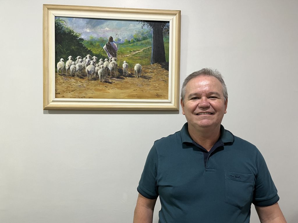 Carnaval: religiões explicam suas visões e posicionamentos sobre o período 3 Um homem sorri na frente de uma pintura retratando um pastor com ovelhas. A pintura está emoldurada e pendurada na parede. Carnaval: religiões explicam suas visões e posicionamentos sobre o período