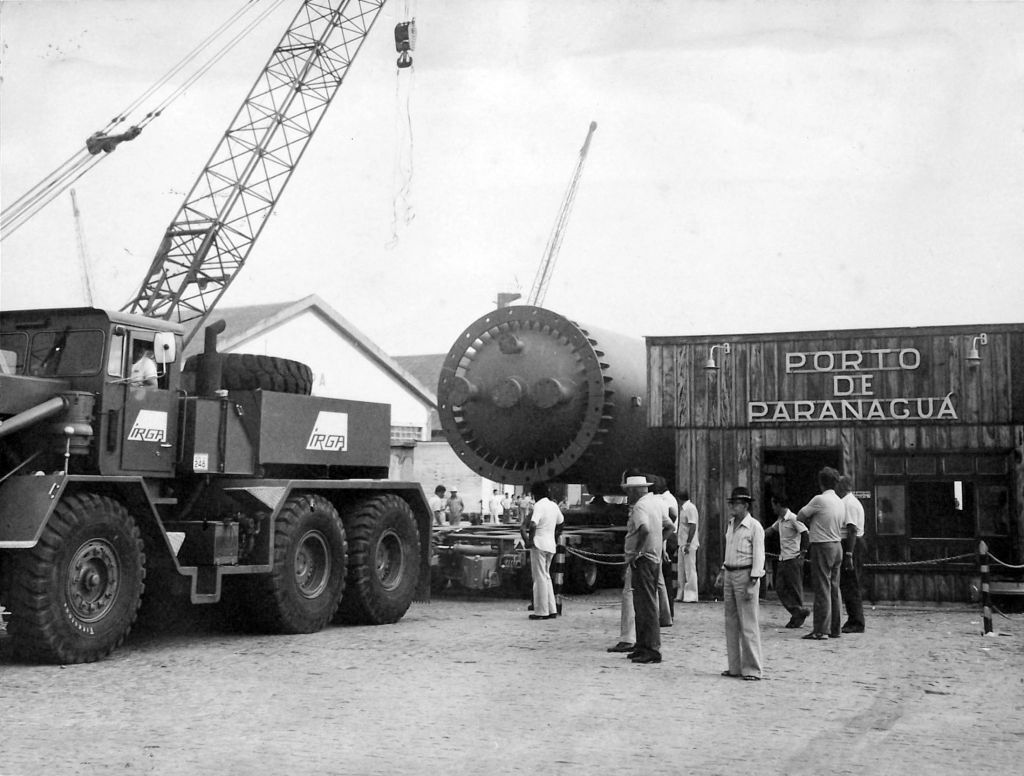 Portos do Paraná: 90 anos de história impulsionando o desenvolvimento de Paranaguá 7 Caminhão guindaste e operários no Porto de Paranaguá, com grande equipamento de carga e armazém ao fundo em imagem preto e branco. Portos do Paraná: 90 anos de história impulsionando o desenvolvimento de Paranaguá