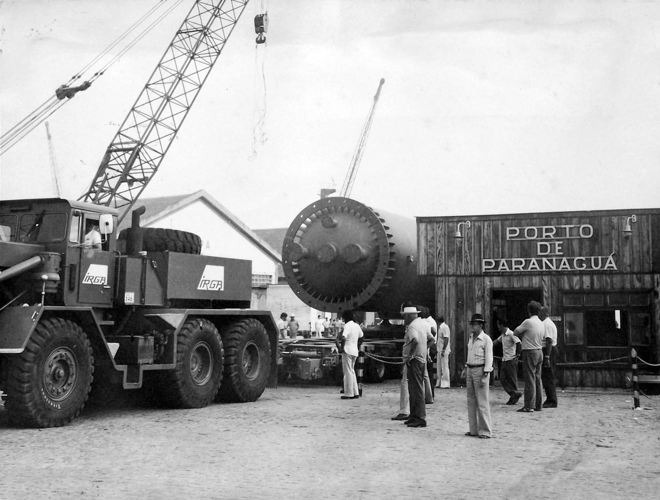 Caminhão guindaste e operários no Porto de Paranaguá, com grande equipamento de carga e armazém ao fundo em imagem preto e branco. Portos do Paraná: 90 anos de história impulsionando o desenvolvimento de Paranaguá