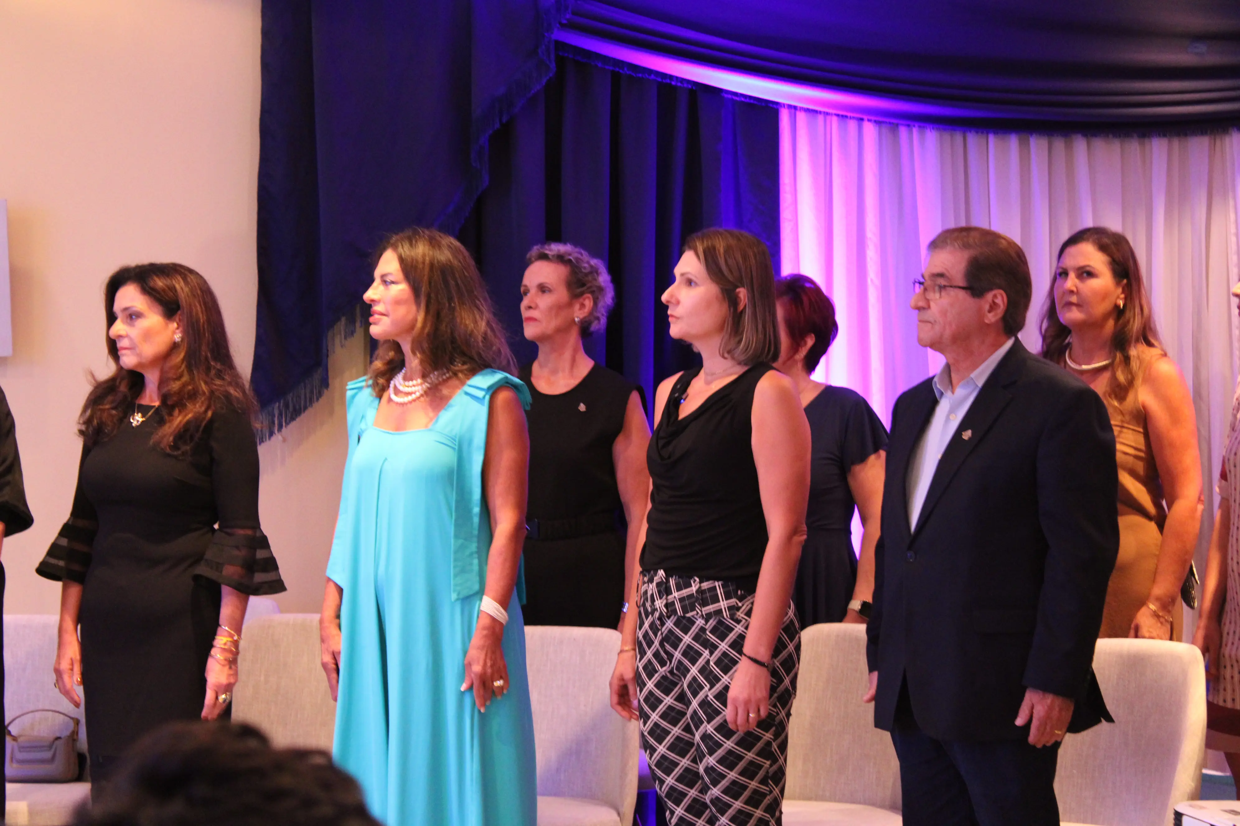 Imagem de um evento social onde várias mulheres e um homem estão em pé, todos com expressão de seriedade. O ambiente é decorado com cores sóbrias e luzes suaves. Palestra “Mulher, empreendedorismo e justiça: construindo caminhos para a autonomia e igualdade” aconteceu na Aciap Paranaguá