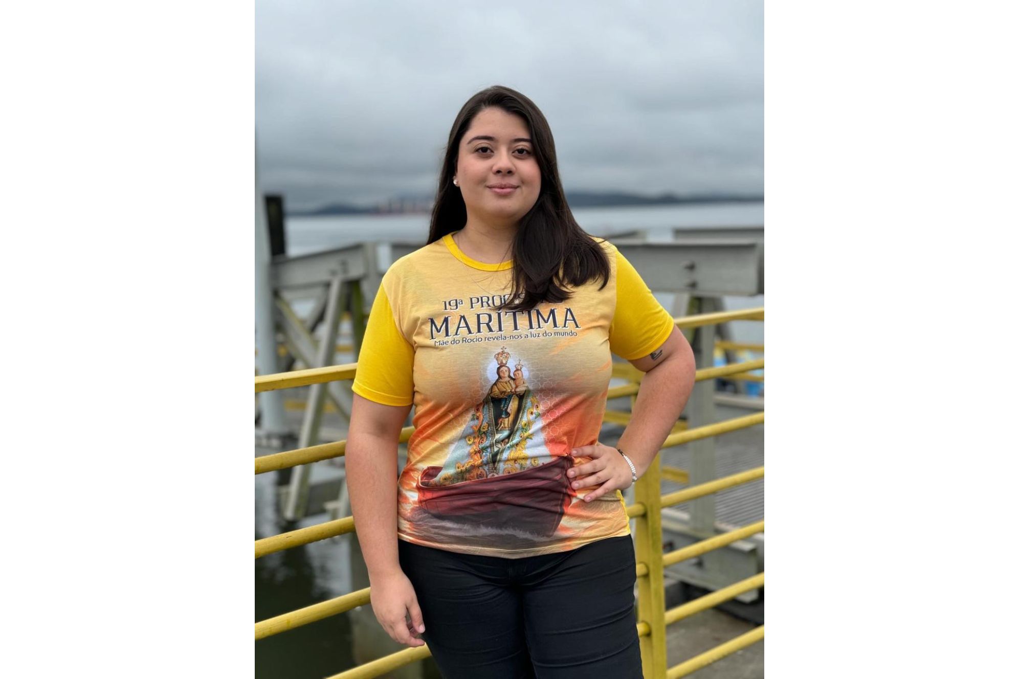 Imagem de mulher usando camiseta amarela com estampa de Nossa Senhora, representando a 19ª Procissão Marítima, em ambiente portuário. Jovem realiza vaquinha para auxiliar no pagamento do tratamento de Degeneração Macular