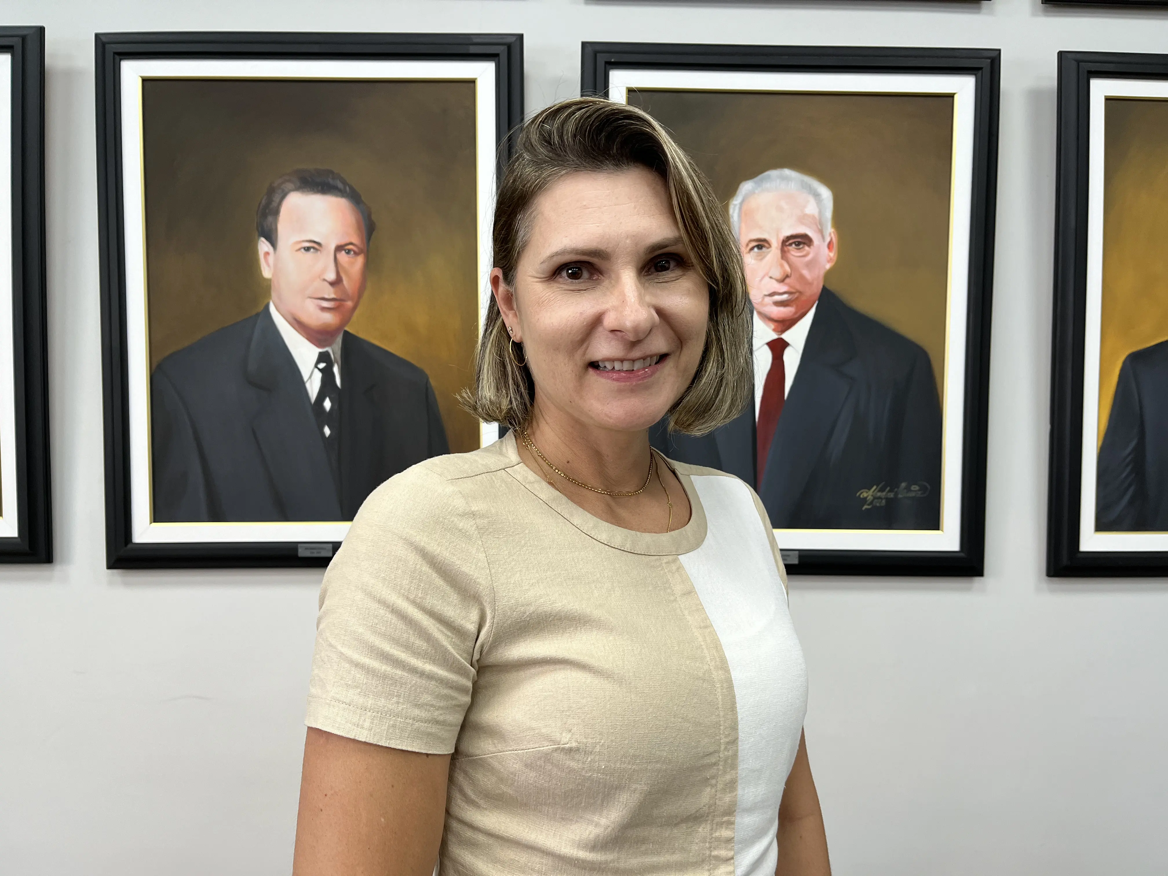 Retrato de mulher sorrindo em frente a quadros de líderes históricos, destacando a importância da memória e história na sociedade. Vice-prefeita de Paranaguá relata sobre a importância da mulher na sociedade