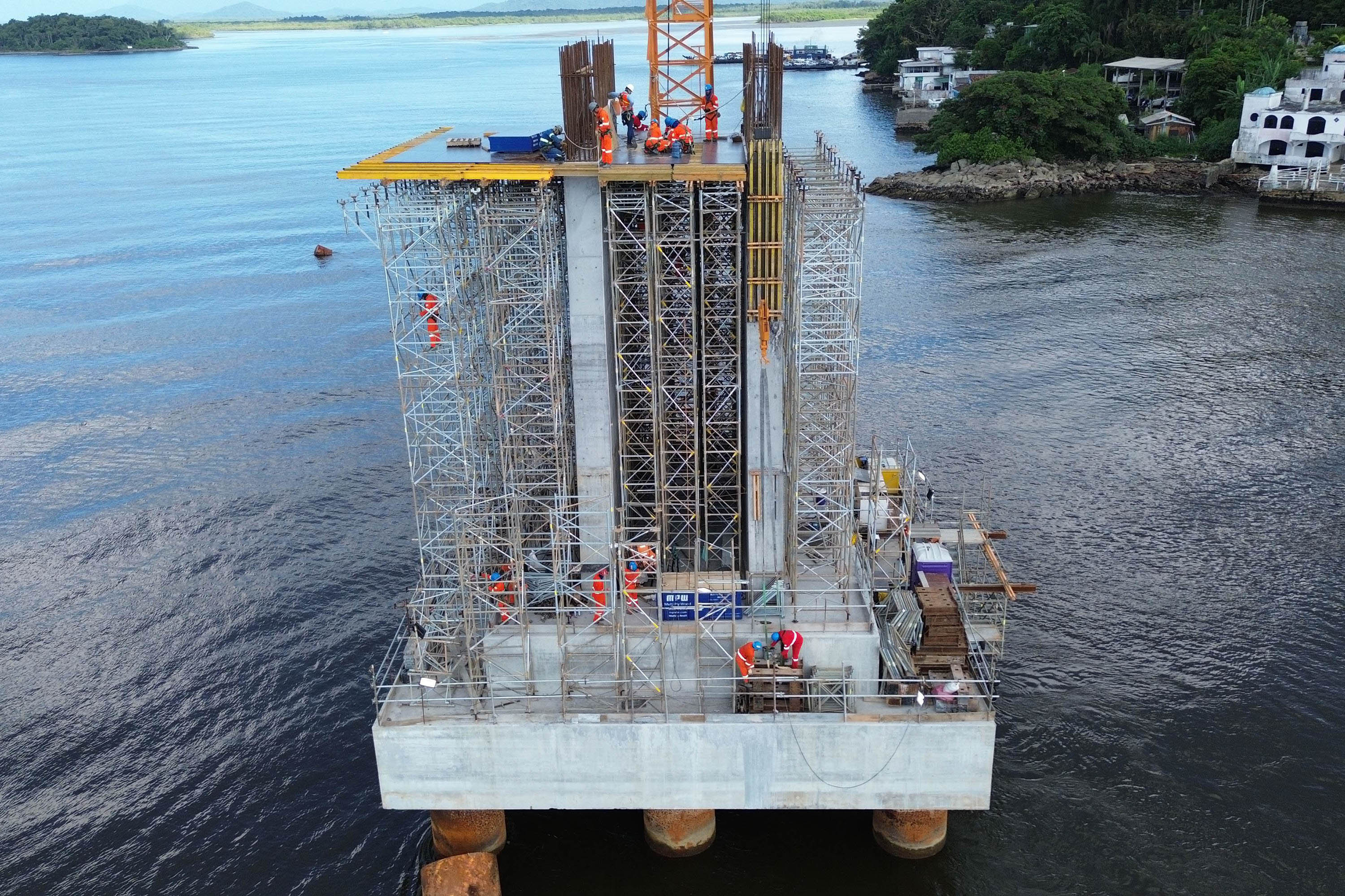 Construção de um pilar em uma plataforma flutuante com trabalhadores em ação. Estrutura de andaime em projeto, situada sobre águas. Com base das torres do trecho estaiado concluída, obra da Ponte de Guaratuba chega a 44%