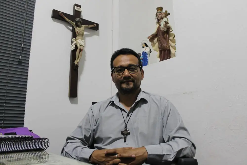 Carnaval: religiões explicam suas visões e posicionamentos sobre o período 2 Imagem de um homem sentado em um escritório com símbolos religiosos ao fundo. Ele usa óculos e um colar com um crucifixo, representando um ambiente de fé e espiritualidade. Carnaval: religiões explicam suas visões e posicionamentos sobre o período