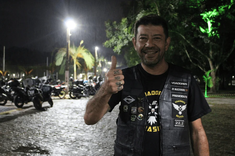 Homem sorridente com colete de motociclista, fazendo sinal de positivo em evento noturno com motocicletas estacionadas ao fundo.