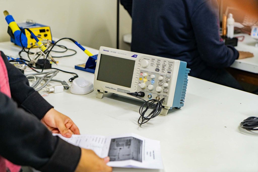 Imagem de um osciloscópio e equipamentos de eletrônica sobre uma mesa de trabalho, com uma pessoa segurando um documento. O osciloscópio é fundamental para medições eletrônicas. Novo recorde: rede estadual tem 50 mil novos alunos na Educação Profissional em 2025