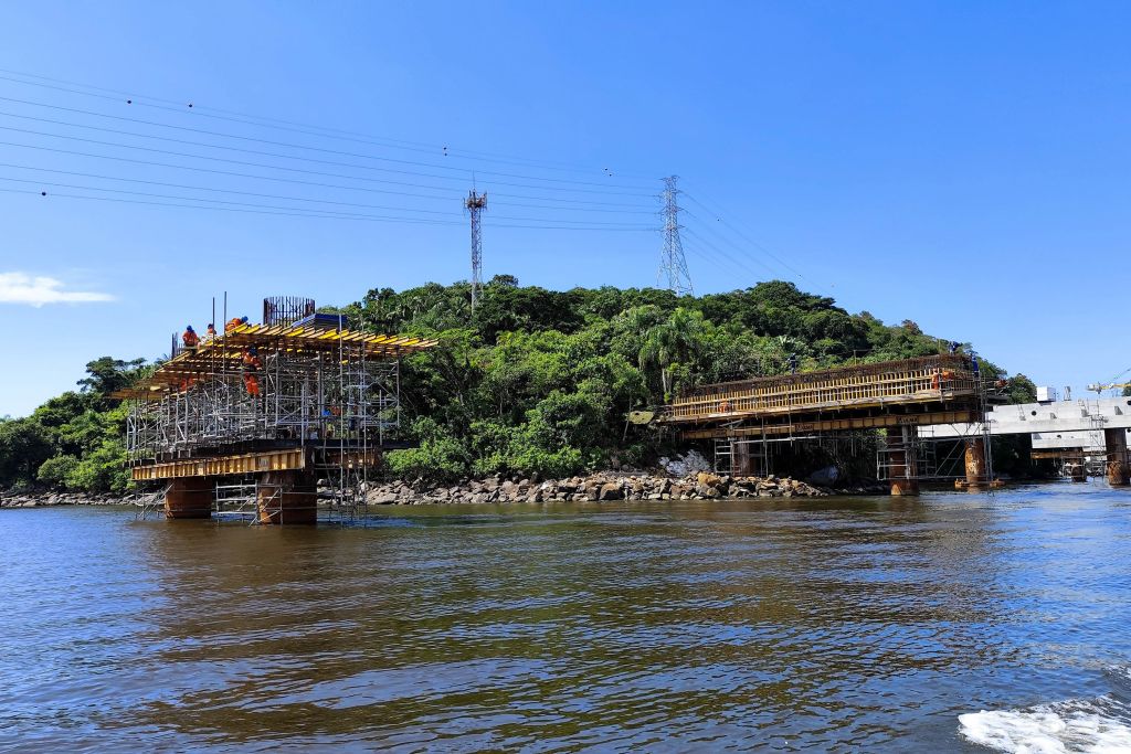 Construção de ponte sobre a água com estruturas de andaime, cercada por vegetação e linhas de energia elétrica ao fundo. Sol radiante e céu claro. Com base das torres do trecho estaiado concluída, obra da Ponte de Guaratuba chega a 44%