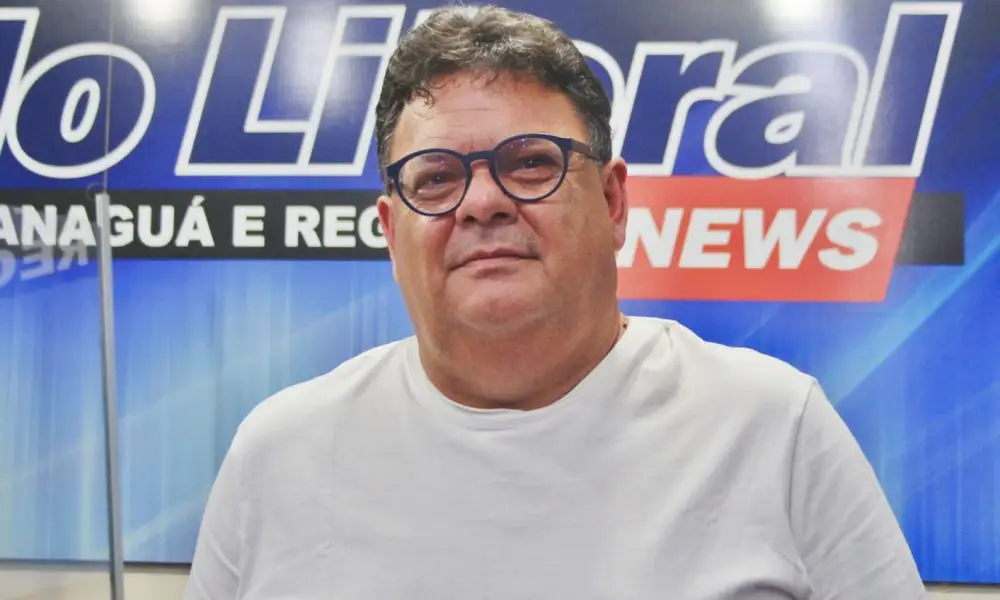 Portos do Paraná: 90 anos de história impulsionando o desenvolvimento de Paranaguá 2 Imagem de um homem sorridente em uma entrevista, com uma camiseta branca, óculos e um fundo que menciona um noticiário. Ele parece confiante e acessível, posando para a foto. Portos do Paraná: 90 anos de história impulsionando o desenvolvimento de Paranaguá