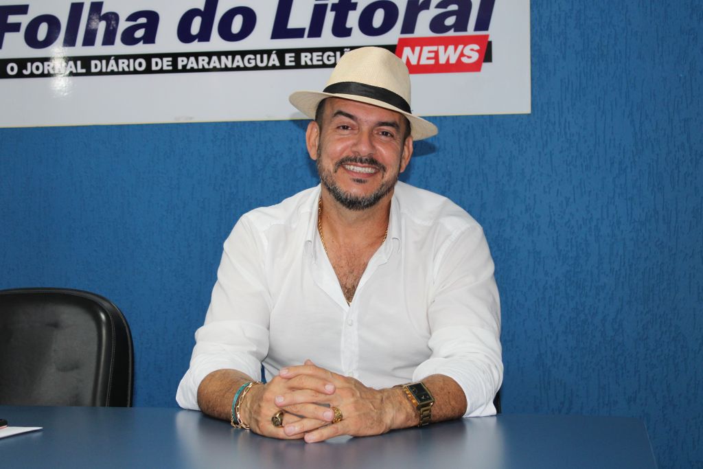 Carnaval: religiões explicam suas visões e posicionamentos sobre o período 5 Imagem de um homem sorridente com um chapéu claro, usando uma camisa branca e sentado à mesa em um ambiente de escritório. O fundo é azul e há uma placa com o nome 'Folha do Litoral'. Carnaval: religiões explicam suas visões e posicionamentos sobre o período