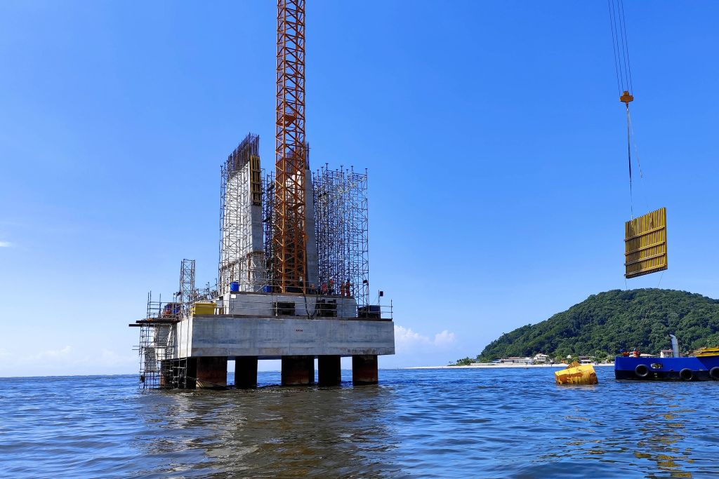 Plataforma em construção sobre a água, com guindaste e estrutura de andaimes, destacando o trabalho de engenharia e construção sustentável. Com base das torres do trecho estaiado concluída, obra da Ponte de Guaratuba chega a 44%