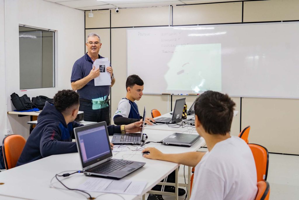 Aula interativa em sala de informática com um professor apresentando conceitos a um grupo de alunos em uma mesa equipada com laptops. Novo recorde: rede estadual tem 50 mil novos alunos na Educação Profissional em 2025