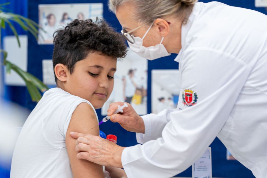 Vacinação começa na segunda-feira nas escolas públicas e privadas 2 Criança recebendo vacina aplicada por profissional de saúde com máscara, em ambiente de vacinação, promovendo a saúde infantil. Vacinação começa na segunda-feira nas escolas públicas e privadas
