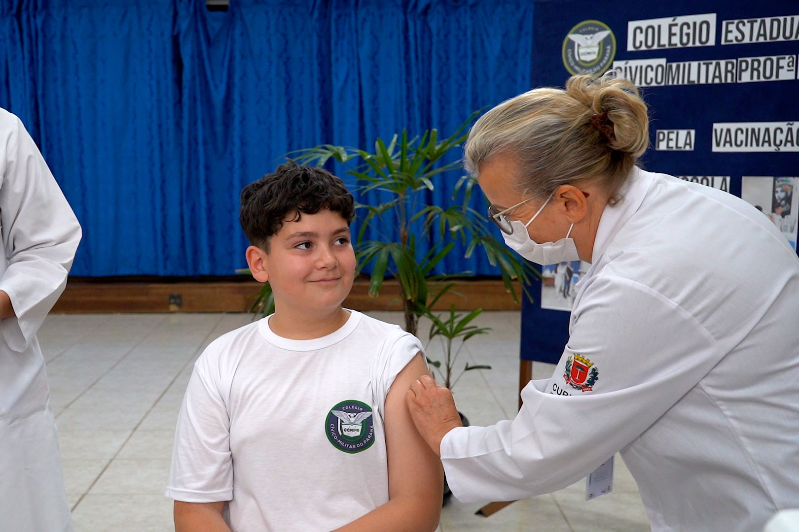 Criança recebendo vacina em colégio durante campanha de vacinação. Mãe e profissional de saúde felizes, simbolizando saúde infantil e prevenção. Vacinação começa na segunda-feira nas escolas públicas e privadas
