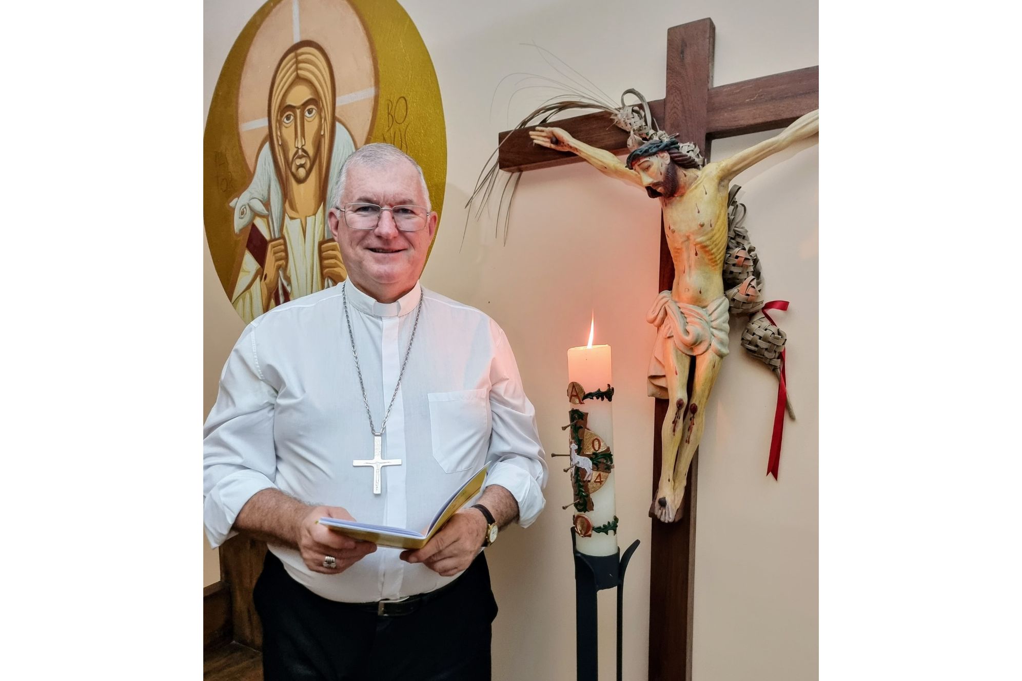 Um sacerdote sorridente posando em frente a um crucifixo e uma vela acesa, com arte religiosa ao fundo, simbolizando a fé e a espiritualidade. Bispo Dom Edmar Peron ressalta importante reflexão para a Páscoa